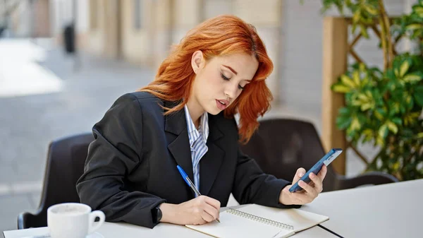 コーヒーショップのテラスでノートを取るスマートフォンを使用して若い赤毛の女性ビジネスワーカー — ストック写真