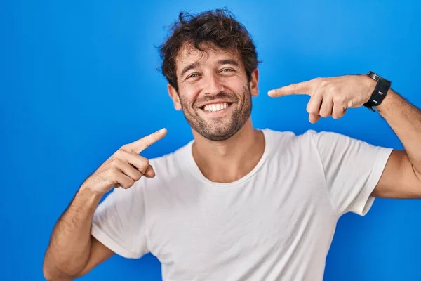 ヒスパニック系の若い男が青の背景に立って陽気に示す笑顔と指の歯と口を指しています 歯の健康の概念 — ストック写真