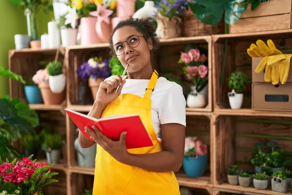 African american woman florist reading notebook with doubt expression at flower shop