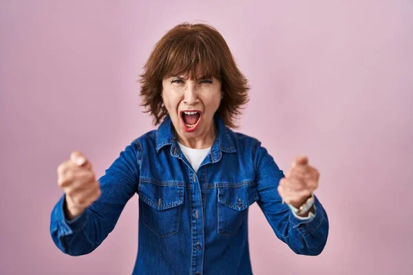 ピンク色の背景で立っている中年女性は怒りと怒りで叫んでいる間に怒りと怒りの拳を上げる 怒りと攻撃的な考え方 — ストック写真