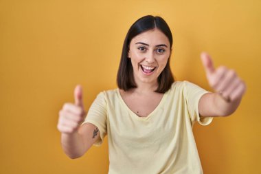 İspanyol kız sarı arka plan üzerine günlük tişört giyip olumlu jestler yapıyor, gülümsüyor ve başarı için seviniyor. kazanan jest. 