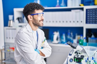 Bilim adamı üniforması giyen genç bir Arap laboratuvarda kollarını kavuşturup oturuyor.