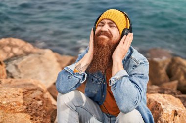 Genç kızıl saçlı, gülümseyen, kendinden emin, deniz kenarında müzik dinleyen bir adam.