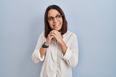 Beyaz arka planda dikilen genç İspanyol kadın. Gergin ve heyecanlı. Elleri çenesinde yan tarafa bakıyor. 