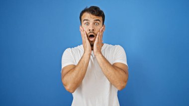 İzole edilmiş mavi arka planda sürpriz bir ifadeyle duran İspanyol genç adam.