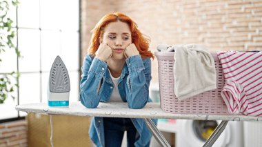 Genç kızıl saçlı kadın çamaşırhanede üzgün bir ifadeyle ütü masasına yaslanıyor.