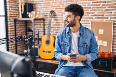 Genç Arap müzisyen, müzik stüdyosunda akıllı telefon kullanarak gülümsüyor.