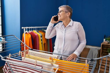 Orta yaşlı bir kadın çamaşır odasında çamaşır ipine elbise asarak akıllı telefondan konuşuyor.