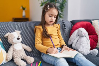 Adorable hispanic girl drawing on notebook sitting on sofa at home