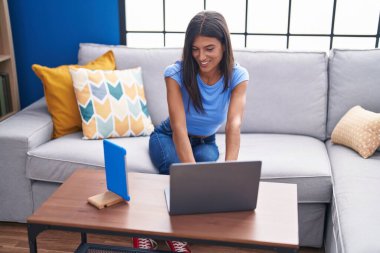 Young beautiful hispanic woman using laptop and touchpad sitting on sofa at home