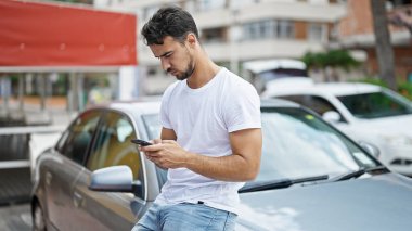 Young hispanic man using smartphone sitting on car at street