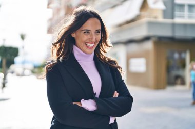 Genç İspanyol kadın gülümsüyor. Kollarını kavuşturup caddede duruyor.