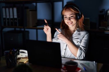 Young caucasian woman working at the office at night smiling and looking at the camera pointing with two hands and fingers to the side. 