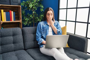 İspanyol kadın evde dizüstü bilgisayar kullanıyor. Ağzını eliyle kapatıyor. Şok geçiriyor ve hata yapmaktan korkuyor. şaşırmış ifade 