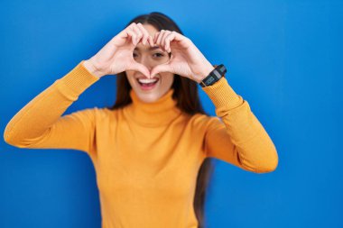 Genç esmer kadın mavi arka planda duruyor. El ve parmaklarıyla kalp şeklinde, tabelanın içinden bakıp gülümsüyor. 