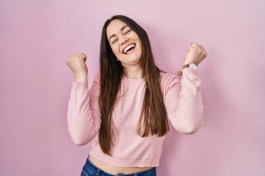 Young brunette woman standing over pink background celebrating surprised and amazed for success with arms raised and eyes closed. winner concept. 