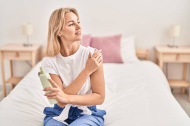 Young blonde woman applying lotion on skin at bedroom