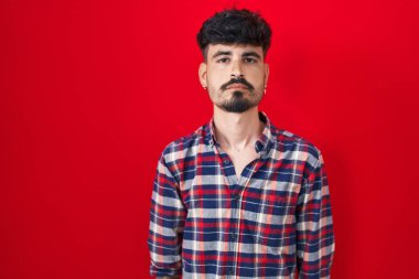 Kırmızı arka planda duran sakallı İspanyol genç adam yüzünde ciddi bir ifadeyle rahatladı. Basit ve doğal bir şekilde kameraya bakıyorsun.. 