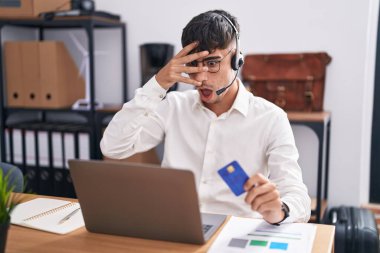 Bilgisayarda çalışan genç İspanyol bir adam elinde bir bilgisayar kartıyla şok içinde yüzünü ve gözlerini kapayarak utangaç ifadesiyle parmaklarından bakıyor.. 