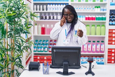 Afro-Amerikalı kadın eczacı elinde ilaç şişesiyle eczanede akıllı telefondan konuşuyor.