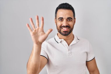 Beyaz arka plan üzerine günlük giysiler giyen, sakallı genç İspanyol adam beş numaralı parmaklarıyla işaret ederken kendinden emin ve mutlu bir şekilde gülümsüyor.. 