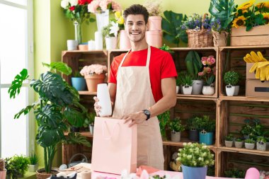 Young hispanic man florist smiling confident putting plant jar on shopping bag at flower shop