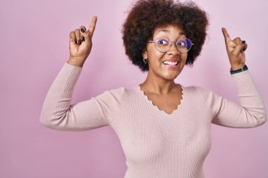 Genç Afrikalı Amerikalı kadın pembe arka planda duruyor. Şaşırmış, şaşırmış ve parmaklarıyla işaret ediyor ve kollarını kaldırıyor.. 