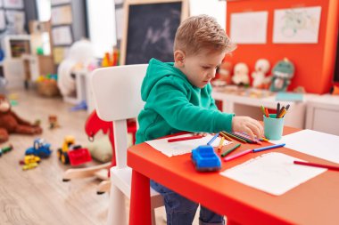 Sevimli beyaz çocuk anaokulunda masa başında oturmuş not defterine resim çiziyor.