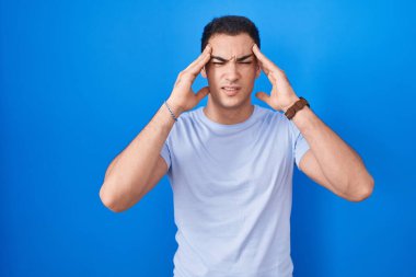 Young hispanic man standing over blue background with hand on head, headache because stress. suffering migraine. 