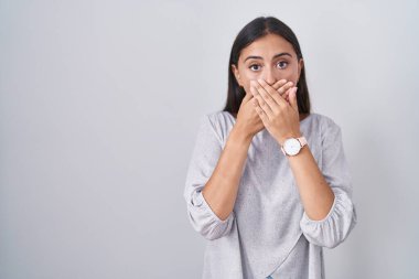 Young hispanic woman standing over white background shocked covering mouth with hands for mistake. secret concept. 