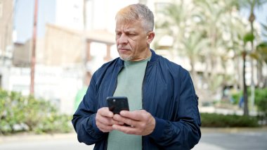 Orta yaşlı, kır saçlı bir adam parkta akıllı telefon kullanıyor.