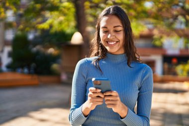 Genç Afrikalı Amerikalı kadın parkta akıllı telefon kullanarak gülümsüyor.