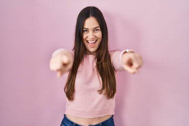 Genç esmer kadın pembe arka planda dikiliyor. Seni ve kamerayı işaret ediyor. Parmaklıyor. 