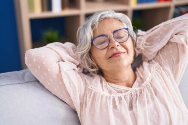 Orta yaşlı, kır saçlı, elleri başının üstünde, evinde kanepede oturan bir kadın.