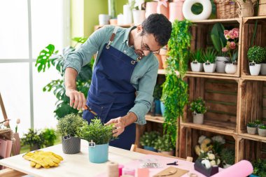 Young hispanic man florist cutting plant at flower shop