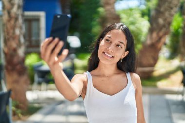 Genç, güzel, İspanyol bir kadın gülümsüyor. Parktaki akıllı telefonun yanında özçekim yapıyor.