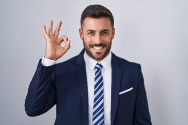 Yakışıklı İspanyol takım elbiseli ve kravatlı adam gülümseyen olumlu el ve parmaklarıyla tamam işareti yapıyor. başarılı ifade. 