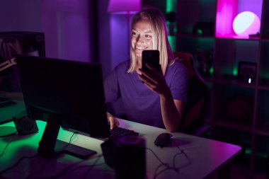 Young blonde woman streamer using computer and smarpthone at gaming room