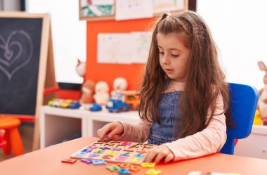 Adorable hispanic girl playing with maths puzzle game sitting on table at kindergarten