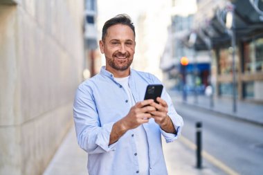 Sokakta akıllı telefon kullanan, kendinden emin gülümseyen beyaz bir genç.