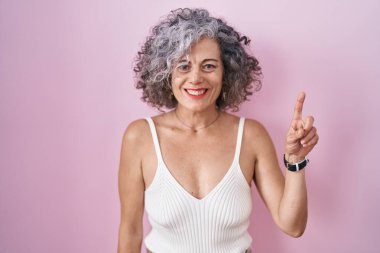 Pembe arka planda duran gri saçlı orta yaşlı kadın bir numaralı parmağı gösterip işaret ederken kendinden emin ve mutlu gülümsüyor.. 
