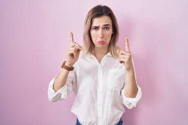 Pembe arka planda duran genç ve güzel bir kadın üzgün ve üzgün görünüyor, parmaklarıyla yön gösteriyor, mutsuz ve depresif.. 