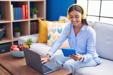 Young beautiful hispanic woman using laptop and touchpad sitting on sofa at home