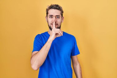 Hispanic man with beard standing over yellow background asking to be quiet with finger on lips. silence and secret concept. 