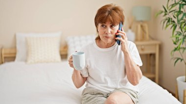 Middle age woman talking on smartphone drinking coffee at bedroom
