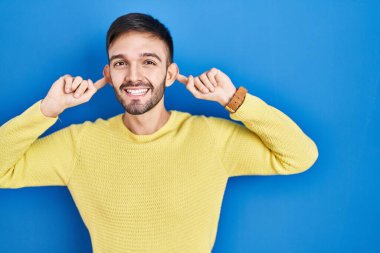 Hispanic man standing over blue background smiling pulling ears with fingers, funny gesture. audition problem 