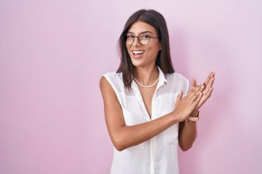 Pembe arka planda duran esmer genç bir kadın gözlükleriyle alkışlıyor ve mutlu ve mutlu, gururlu elleri alkışlıyor. 