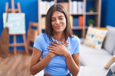 Esmer, genç bir kadın evdeki kanepede oturuyor elleri göğsünde, gözleri kapalı ve yüzünde minnet dolu bir jest ile gülümsüyor. sağlık kavramı. 