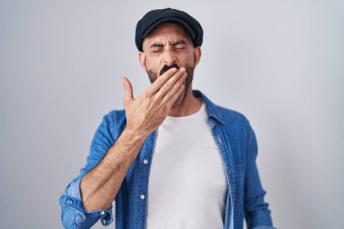 Hispanic man with beard standing over isolated background bored yawning tired covering mouth with hand. restless and sleepiness. 