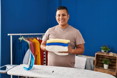 Ütü yaptıktan sonra katlanmış çamaşırları tutan İspanyol genç adam mutlu ve havalı bir gülümsemeyle gülümsedi. diş gösterme. 